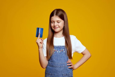 Gülümseyen çocuk mavi bir kredi kartı tutuyor. Parlak sarı bir arkaplan önünde merak ve heyecan dolu. Finansal eğitim içeriği ve çocuklarla ilgili reklamlar için ideal.