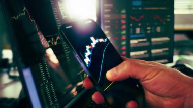 Şifreli Para Birimi Akıllı Telefon Engelleme Teknolojisi Finans Konsepti. Ekranda Bitcoin 'in şamdan çizelgesiyle el ele tutuşan akıllı telefon, finansal piyasa verilerini gösteren birden fazla monitörle çevrili.