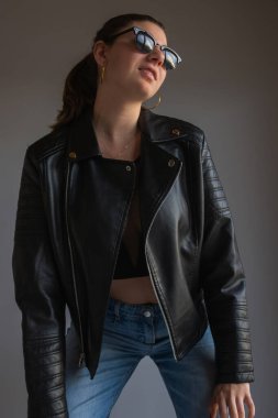 Deri ceket ve güneş gözlüğü takan şık, genç bir kadın. Moda kampanyaları, sokak kıyafetleri reklamları ve yaşam tarzı reklamları için mükemmel..