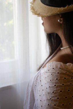 Hasır şapkalı ve beyaz gözlüklü genç bir kadın pencereden dışarı bakıyor, yumuşak doğal ışıkla yıkanmış, sakin ve düşünceli bir anın tadını çıkarıyor.