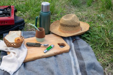 Kırsal bir pano ve battaniyede piknik malzemeleri, taze ekmek, geleneksel tarba eşi ve termos, kırsal alanda rahat bir yemek için hasır şapka ve kompakt kamp malzemeleri.