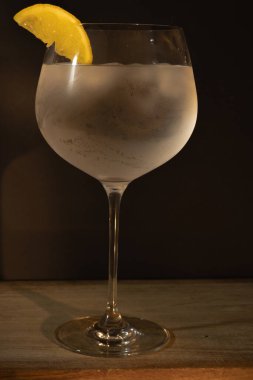 Soğuk cin ve tonik kokteyli buzla soğutulmuş, canlı bir limon dilimiyle süslenmiş, koyu arkaplanlı sofistike bir bardakta canlandırıcı alkollü içecek sergileniyor.