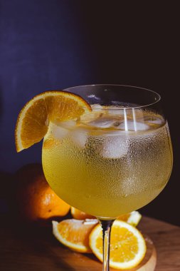 Taze portakal dilimi ve erimiş buzlu cin tonik, camda yoğunlaşma, karanlık bir stüdyonun arka planında soğuk, turuncu bir yaz kokteyli, yakın çekim ve davetkar