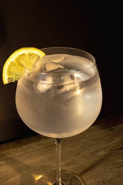 Buzlu bardakta buzlu ve limonlu cin ve tonik tazeleniyor. Yoğunlaşma, kırsal ahşap bir masada çarpıcı bir ışık altında parıldıyor. Soğuk ve zarif bir içecek.