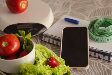 Taze domates ve marul, bir mutfak terazisi, mezura, not defteri ve akıllı telefon bir masanın üzerinde, beslenme düzenine, sağlıklı yaşama ve ağırlık yönetimine yapılandırılmış bir yaklaşımı temsil ediyor.