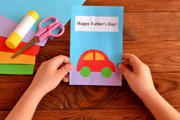 Child holds a greeting card in his hands. Kids crafts for Father's day. Card with message Happy Father's day. Holiday greeting card gift for dad from child. Colored paper sheets, scissors, glue stick. Wooden background