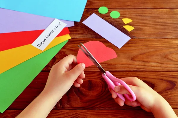 El niño hace un regalo para papá. El niño mantiene las tijeras en las manos y corta un coche de papel. Hojas de papel de colores, tijeras, detalles recortados. Idea de regalo hecha en casa del día del padre. Fondo de madera. Lugar de trabajo infantil — Foto de Stock