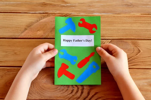 Child holds Fathers day greeting card gift in his hands. Homemade Fathers day greeting card idea. Preschool and kindergarten paper tools collage crafts. Handmade card idea. Happy Father's day. Wooden table — Stock Photo, Image