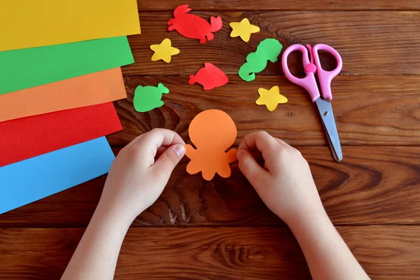 Découpez des animaux marins en papier coloré - poulpe, poisson, étoile de mer, hippocampe, crabe. Le petit enfant tient dans ses mains une pieuvre artisanale. Enfants bricolage, feuilles de papier coloré, ciseaux sur fond en bois — Photo