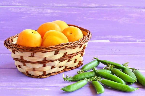 Verse rijpe abrikozen in een mand en peulen van groene erwten op paarse houten achtergrond. Gezond en smakelijk eten. Macro — Stockfoto