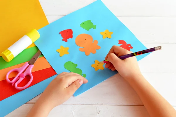 El niño sostiene un lápiz y dibuja. Niño haciendo una tarjeta con animales marinos y peces. Hojas de papel de colores, tijeras, pegamento, set para niños arte — Foto de Stock