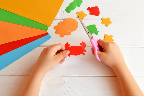 El niño sostiene una tijera y corta cangrejo de papel. Clase de arte en el jardín de infantes. Animales de mar de papel: pulpo, peces, estrellas de mar, caballitos de mar, cangrejos. Artesanía infantil — Foto de Stock