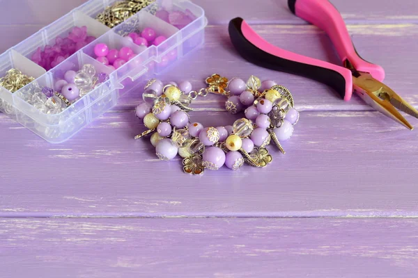 Beautiful bracelet with lilac and white plastic beads, metal flowers and leaves. Organizer with different beads, steel decorative pendants and rings. Pliers. Homemade jewelry concept — Stock Photo, Image