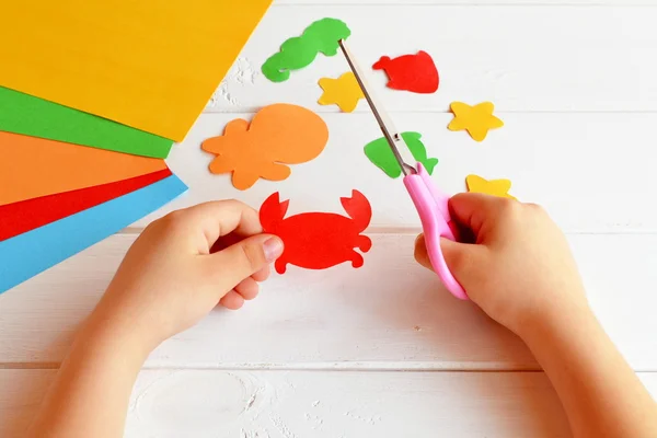 El niño sostiene una tijera y corta cangrejo de papel. Clase de arte en el jardín de infantes. Animales de mar de papel: pulpo, peces, estrellas de mar, caballitos de mar, cangrejos. Artesanía infantil — Foto de Stock