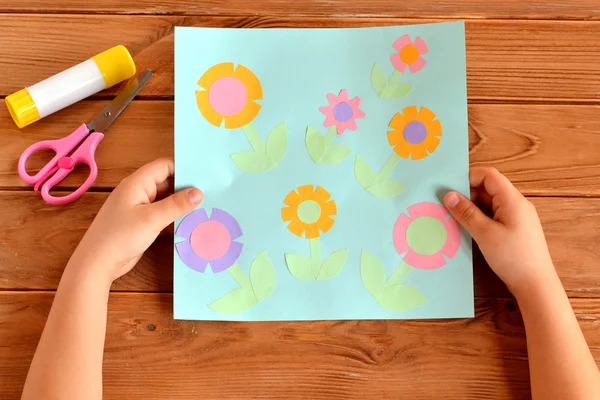 Il bambino tiene in mano una carta con dei fiori. Colla, forbici su un tavolo di legno. Progetto di arte infantile — Foto Stock