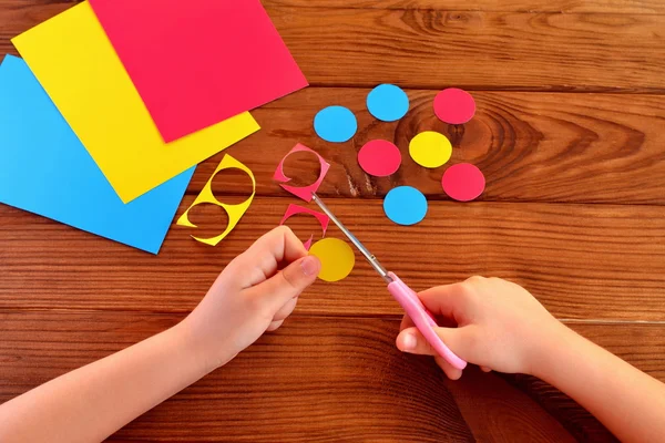 El niño sostiene tijeras y papel y corta el círculo. Hojas de papel de colores. Fondo de madera marrón — Foto de Stock