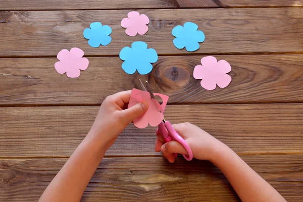 Το παιδί κόβει ένα λουλούδι από χαρτί. Το παιδί κρατάει ψαλίδι και χαρτί στα χέρια του. Χαρτολουλούδια σε ξύλινο τραπέζι. Θερινές δραστηριότητες για παιδιά σε οικιακές χειροτεχνίες. Δραστηριότητες κοπής για παιδιά πρακτική ψαλιδιού — Φωτογραφία Αρχείου