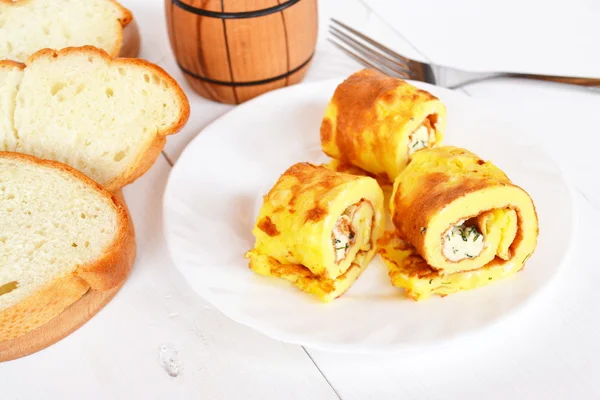 Омлетні рулети з сиром і зеленню на тарілці. Шматочки хліба на обробній дошці, виделка на білому дерев'яному фоні. концепція сніданку — стокове фото