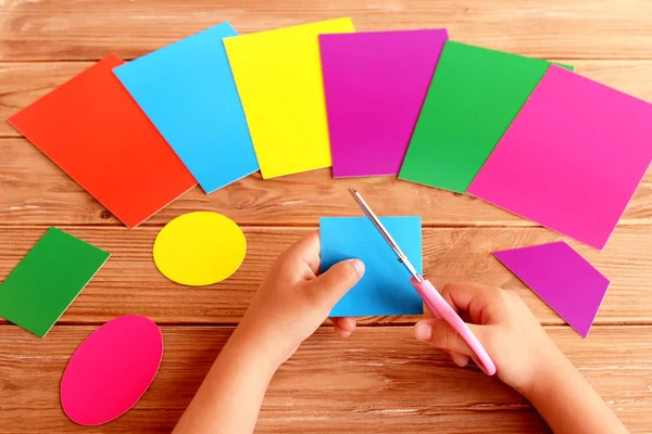 Niño sostiene tijeras y cartón cuadrado en sus manos. Hojas de cartón de colores y diferentes figuras geométricas en una mesa de madera. El niño aprende a cortar formas de papel. Desarrollo de la primera infancia — Foto de Stock