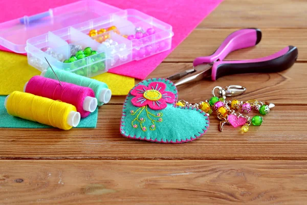Beautiful keychain felt heart with colorful beads and flower. Box of beads, tools, sewing set, felt sheets on a wooden table. Children fabric project — Stock Photo, Image