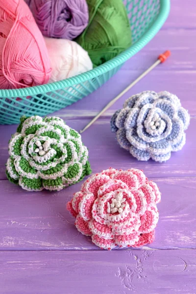 Pink, green and blue crochet flowers decorated with beads. Cotton yarn skeins in basket, hook and bright knitted roses on lilac wooden background — Stock Photo, Image