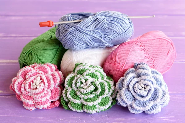 Pink, green and blue crochet roses decorated with beads. Cotton yarn skeins, hook and bright knitted flowers on wooden background lilac — Stock Photo, Image