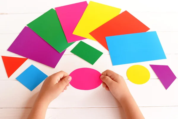 Niño sostiene cartón ovalado rosa en las manos. El niño aprende colores y formas geométricas. Alcanza el concepto. Diferentes tarjetas de cartón en una mesa — Foto de Stock