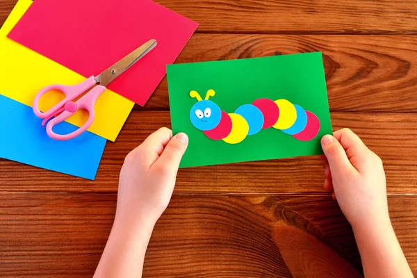 Barn händer som håller papper applique caterpillar. Ark, sax på brun trä bakgrund — Stockfoto