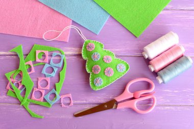 Green Christmas tree decoration toy with colorful balls, scissors, felt scraps, thread set, felt sheets on lilac wooden background. Christmas DIY background. Children winter crafts project. Sewing concept. Christmas tree hand made