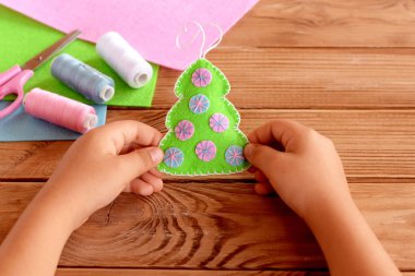 Child holds a felt Christmas tree toy in his hands. Green felt fur tree decorated with pink and blue balls. Christmas crafts diy project for kids. Felt sheets, scissors, thread on wooden background. Felt Christmas tree decortion step by step