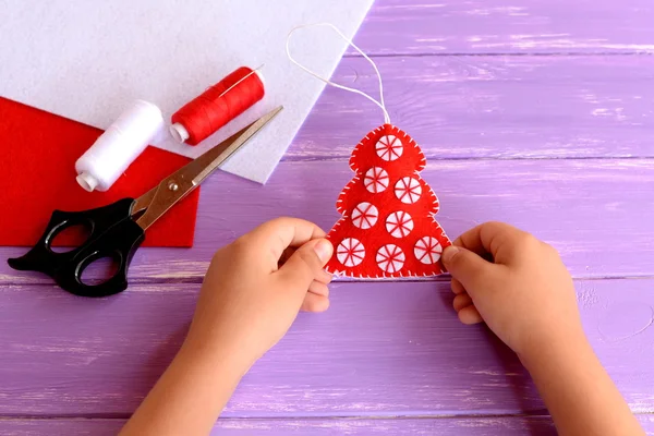 El niño sostiene un adorno de árbol de Navidad de fieltro rojo en sus manos. Árbol de piel de fieltro rojo decorado con bolas blancas. Idea de manualidades navideñas para niños. Hojas de fieltro, tijeras, hilo sobre fondo de madera — Foto de Stock