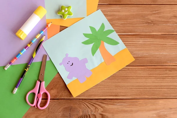 Paper hippo and palm tree applique, colored paper sheets, scissors, pencils, glue, eraser on wooden background with blank space for text. Funny children background. Art and craft project for kids — Stock Photo, Image
