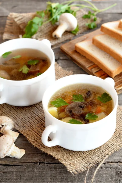 Hem svamp soppa i en skål, vete brödskivor på en skärbräda, färsk rå agaricus och grön persilja på ett träbord. Grönsakssoppa med champinjoner recept. Närbild — Stockfoto