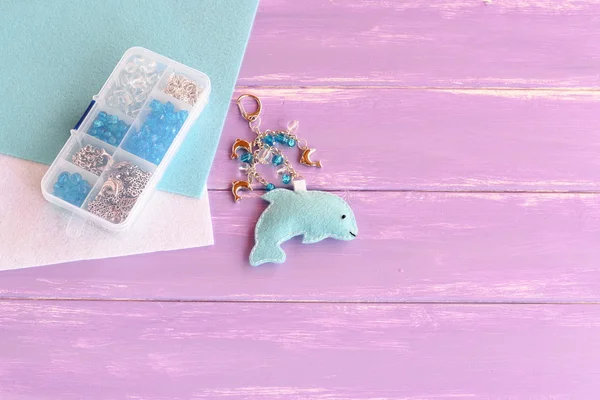 Hand cute felt dolphin keychain. Handbag charms. Handmade car keyring. Set of beads in a plastic box, felt sheets on lilac wooden background with free place for text. Summer crafts for kids concept — Stock Photo, Image