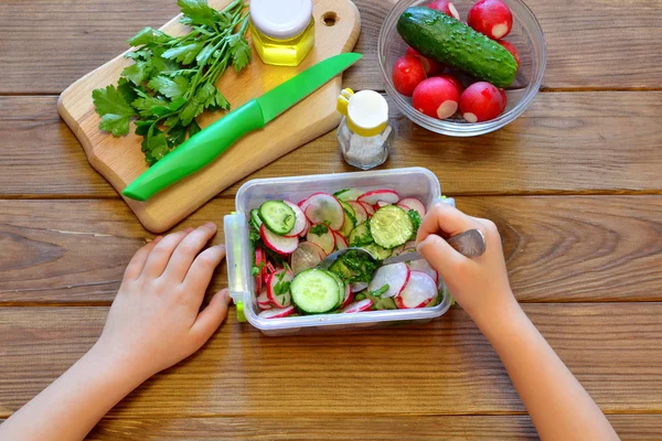 A criança come uma salada com rabanete e pepinos. Conjunto de produtos para cozinhar salada de legumes em fundo marrom de madeira — Fotografia de Stock