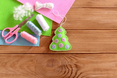 Yeşil keçeli Noel ağacı, pembe ve mavi toplar, iplik, makas, iğne, ahşap masa üzerinde keçeli çarşaf yazı için boş alan. Çocukların yapması kolay bir Noel ağacı zanaatı. Noel süsü arkaplanı. Dikiş malzemeleri.