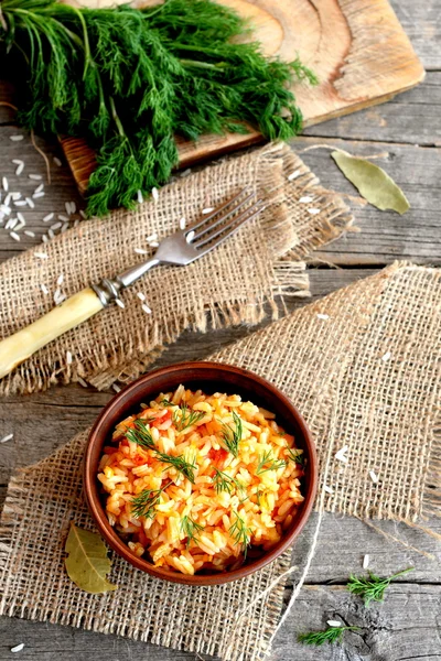 Risotto mit Gemüse in einer Tonschüssel, Gabel, Dillzweigen, einem Schneidebrett auf einem Holztisch. Reis mit Tomaten, Knoblauch und Karotten gekocht und mit Dill garniert. Ansicht von oben — Stockfoto