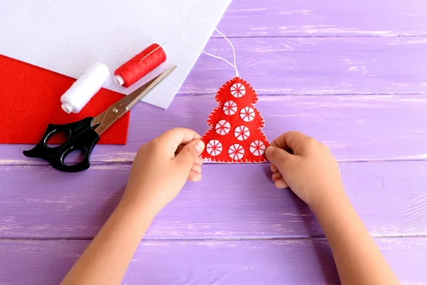 Kind houdt kerstboom ornament in zijn handen. Rood vilt Nieuwjaar kerstboom ornament met witte ballen, schaar, draad, naald, vilt lakens op lila houten achtergrond. Kerst naaien idee — Stockfoto