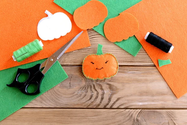 Halloween mini pumpkin decor. Orange felt pumpkin toy, scissors, flat pieces of felt, green and black thread, needle, paper template on wood background. Halloween sewing crafts — Stock Photo, Image