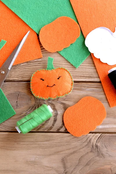 Pretty orange pumpkin ornament with eyes and mouth. Halloween pumpkin made of felt, green thread, scissors, flat pieces of orange and green felt on wood background. Halloween crafts for kids. Top view — Stock Photo, Image