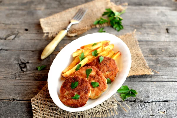 Las croquetas de carne y las patatas fritas en el plato, el tenedor, la rama del perejil y la arpillera sobre el fondo viejo de madera. Patatas hechas con filetes de pavo y fritas en aceite de oliva — Foto de Stock