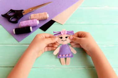 Çocuk bir keçe Bebek elinde tutar ve gösterir. Makas, iş parçacığı, bir tabloda yaprak hissettim. Sevimli doldurulmuş oyuncak el ile yapılır. Çocuklar için kolay dikiş proje 