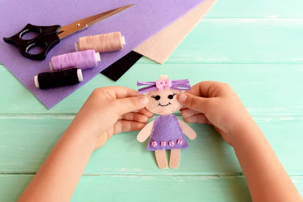 Çocuk bir keçe Bebek elinde tutar ve gösterir. Makas, iş parçacığı, bir tabloda yaprak hissettim. Sevimli doldurulmuş oyuncak el ile yapılır. Çocuklar için kolay dikiş proje 