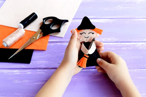 Niño sostiene una muñeca bruja de Halloween en sus manos y lo muestra. Tijeras, hilo, aguja, fieltro en una mesa de madera. Bruja creativa DIY para niños. Proyecto de decoración para el hogar. Halloween artesanía inspiración —  Fotos de Stock