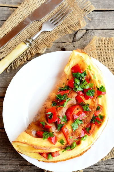 Tortilla de huevo rellena con verduras en un plato. Tortilla vegetariana con pimiento rojo frito, perejil fresco y especias. Tenedor, cuchillo sobre una arpillera y fondo de madera vieja. Receta de desayuno saludable. Vista superior — Foto de Stock