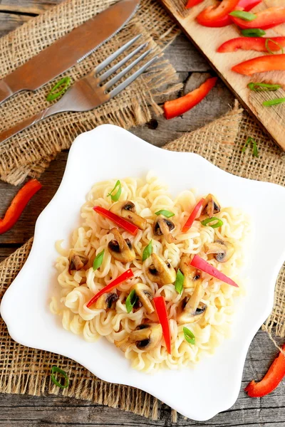 Pasta con champiñones fritos, pimienta roja y cebolla verde. Pasta en un plato, tenedor y cuchillo sobre fondo de madera vieja, pimiento rojo picado y cebollas verdes en una tabla de cortar. Receta vegetariana. Vista superior — Foto de Stock