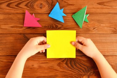 Çocuk elinde bir kağıt kare tutar. Çocuk yapımı origami balığı. Adım. Ahşap bir masa üzerinde origami balık seti 
