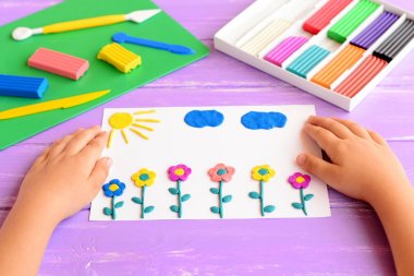 Child shows a card with plasticine flowers, sun and clouds. Supplies for children art crafts on a wooden table. Modeling clay crafts idea for kids. Activity in kindergarten and at home  clipart