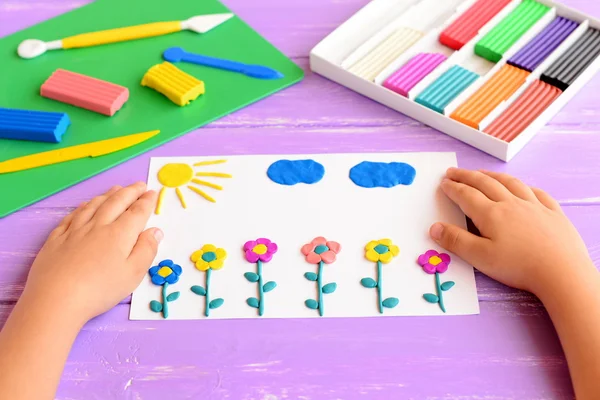 El niño muestra una tarjeta con flores de plastilina, sol y nubes. Suministros para artesanías infantiles en una mesa de madera. Modelar la idea de artesanía de arcilla para niños. Actividad en el jardín de infantes y en casa — Foto de Stock
