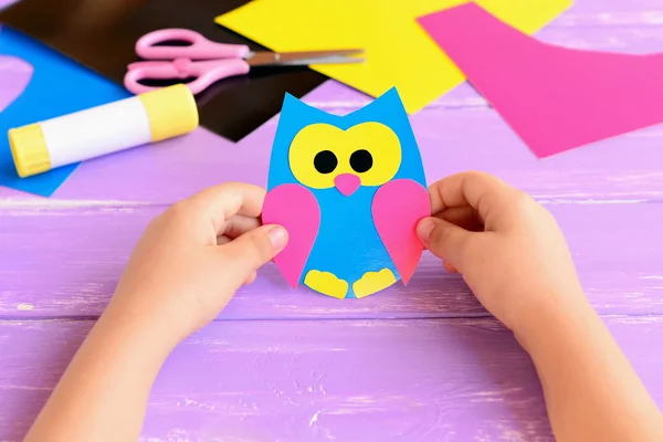 Small child holds paper owl in his hands. Child shows owl crafts. Colored paper sheets, scissors, glue stick on a lilac wooden background. Kids paper crafts. Kids art in kindergarten and at home — Stock Photo, Image